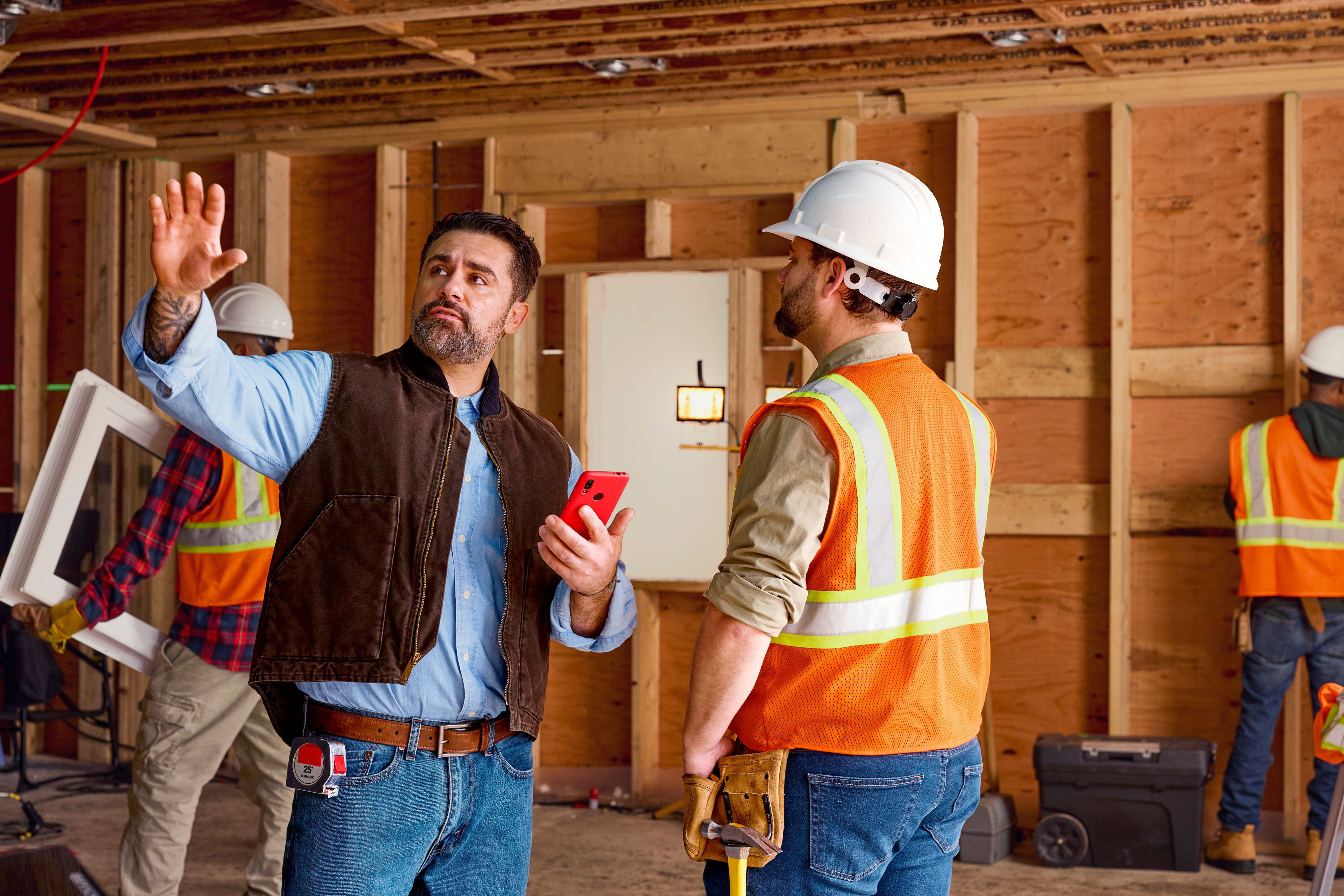 A construction manager holding a smartphone gestures while speaking to a worker wearing a hard hat and safety vest at an indoor construction site.